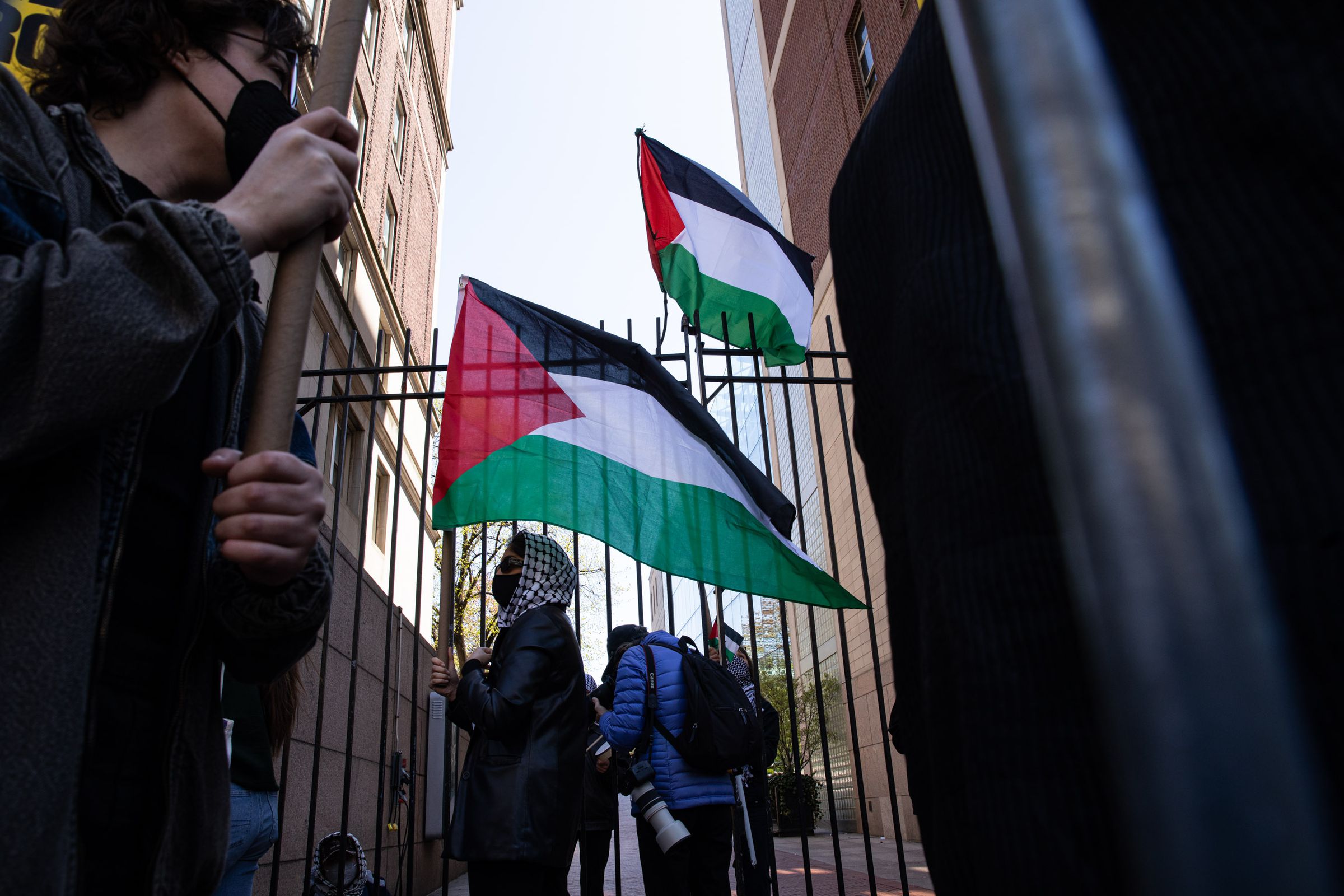 A student wears a mask and kaffiyeh and carries ⁤a Palestinian​ flag.