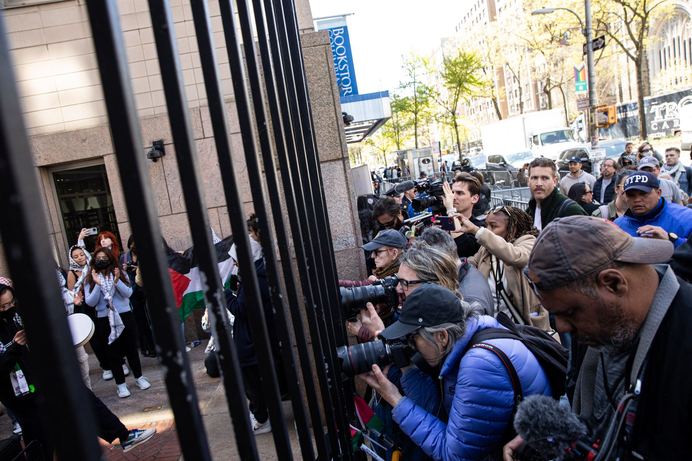 A crowd of photographers presses⁤ in at the wrought-iron⁢ gates of ‍the university. Behind the fence, ‌students demonstrate wearing kaffiyehs ⁢and waving Palestinian flags.