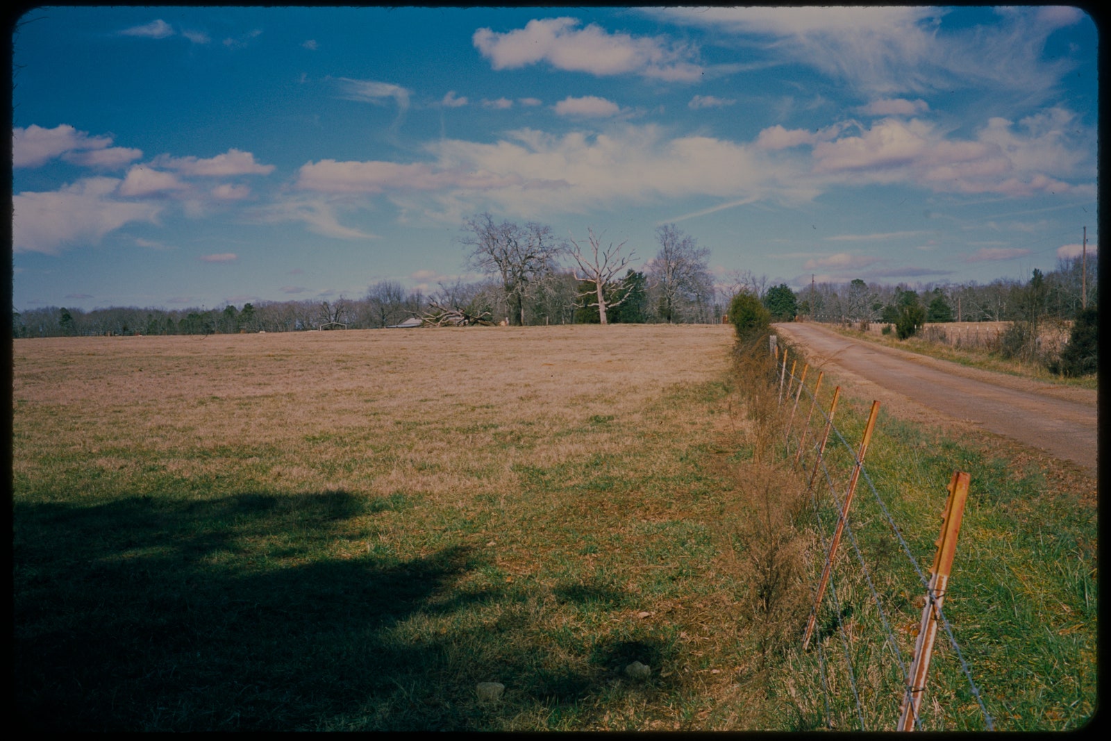 Grainy color photograph of a field of dry grass and a leaning wire fence on the right with a partly cloudy sky in the...